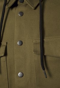 Acercamiento de una chaqueta de color verde oliva con botones metálicos a presión, cordones trenzados negros y bolsillos con solapa con costuras detalladas.