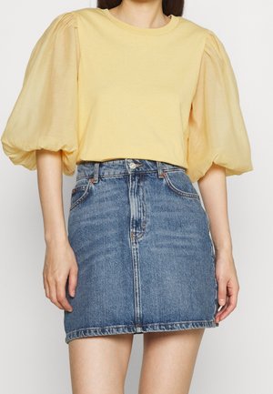Kobieta w żółtej bluzce z puffami, połączonej z niebieską dżinsową spódniczką mini, pokazana od ramion do połowy uda.