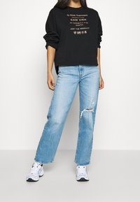 Felpa nera con scritte dorate, jeans blu usurati con vestibilità a gamba larga e sneakers bianche con dettagli neri e rossi.