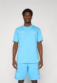 Light blue cotton t-shirt with short sleeves and round neckline, featuring white logos on the chest and left sleeve, paired with matching shorts.