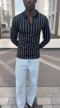 Juodas ilgomis rankovėmis marškinys su baltais vertikaliais dryžiais, su užtrauktuku ir derinamas su šviesiai mėlynomis džinsomis bei juodais sportbačiais. Fone matoma moderni architektūra.