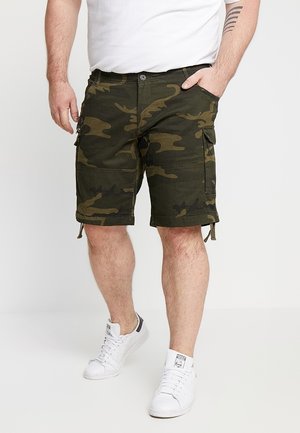 Uomo che indossa pantaloncini cargo mimetici verdi, sneakers bianche e una t-shirt bianca, in piedi con una mano in tasca.