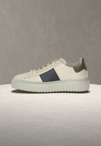 Baskets crème avec semelle épaisse côtelée, panneau latéral bleu marine, patch de talon olive et lacets crème sur fond neutre.