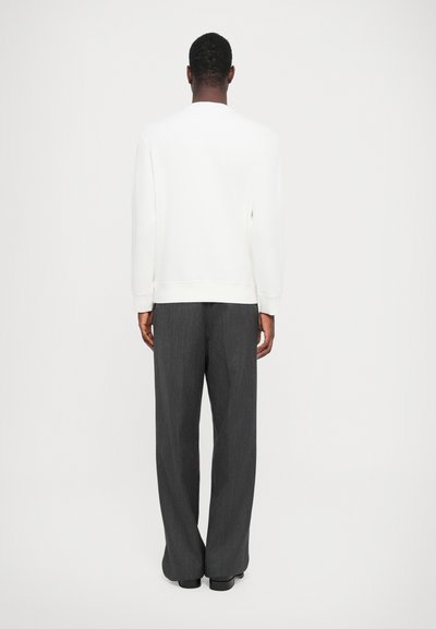 Sudadera blanca con puños y dobladillo acanalados, combinada con pantalones de rayas gris oscuro de corte ancho, de pie frente a un fondo neutro.