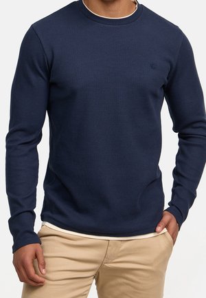 Homme portant une chemise à manches longues texturée bleu foncé et un pantalon beige, une main dans la poche, sur un fond uni.