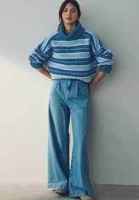 Übergroßer blauer und hellblauer gestreifter Strickpullover mit hohem Kragen, kombiniert mit hochgeschnittenen weit geschnittenen Jeans.