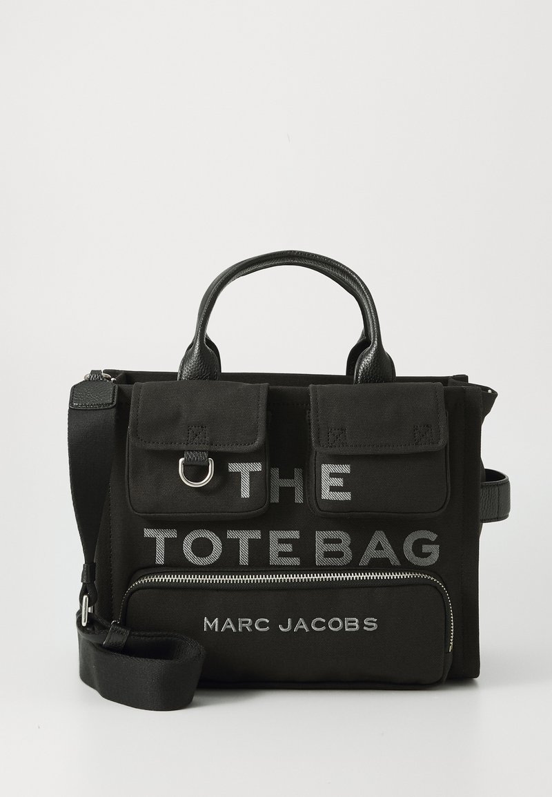 Black canvas tote bag featuring "THE TOTE BAG" and "MARC JACOBS" in bold white lettering, with a zip pocket and dual top handles.