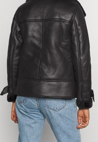 Personne portant un jean bleu clair et une veste en cuir avec doublure en shearling noire, dotée de sangles à boucles sur les poignets, vue de dos.