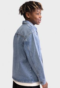 Light blue denim jacket with a classic design, featuring a collar, side pockets, and button closures. Textured fabric with subtle wash effect.