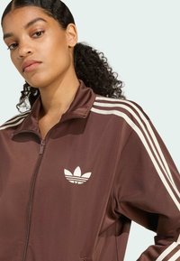 Veste zippée marron en tissu lisse, avec des accents blancs à trois rayures sur les épaules et un logo blanc sur la poitrine.