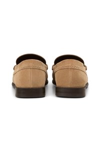 Beige Wildlederslipper mit einem niedrigen braunen Absatz, featuring einen abgerundeten Rücken und subtilen Logo-Details auf dem Obermaterial. Glatte Textur durchgehend.
