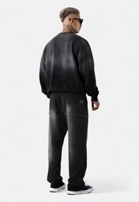 Schwarzer Sweatshirt mit verlaufendem Finish und lockerer Passform, kombiniert mit dunklen Hosen mit genähten Taschen. Getragen mit schwarzen Sneakers und Sonnenbrille.