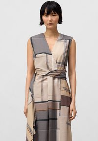 Robe sans manches en tissu texturé présentant un imprimé géométrique beige, gris et marron, avec une taille nouée pour mettre en valeur la silhouette.