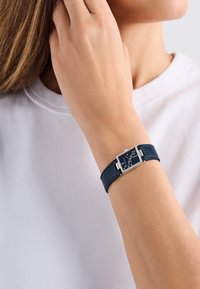 Kék bőr övvel ellátott óra, téglalap alakú ezüst tokból és sötétkék számlapból áll, fehér órajelzőkkel és ezüst kiegészítőkkel.