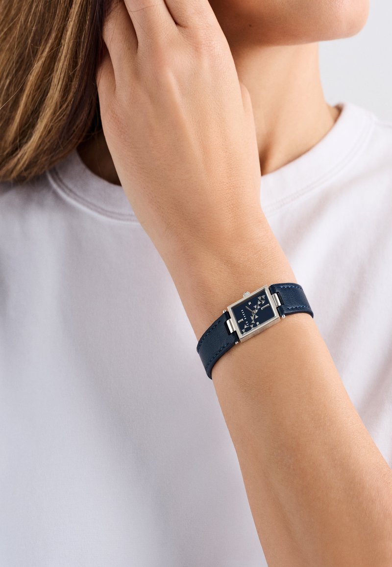 Kék bőr övvel ellátott óra, téglalap alakú ezüst tokból és sötétkék számlapból áll, fehér órajelzőkkel és ezüst kiegészítőkkel.