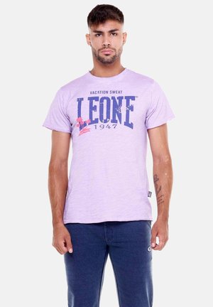 Man staat frontaal, draagt een lichtpaarse "Leone 1947" t-shirt en donkerblauwe broek met een zichtbare tatoeage op de onderarm.