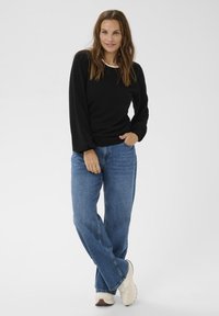 Kvinde med langt brunt hår iført en sort sweater, blå jeans og hvide sneakers, stående med den ene hånd i lommen og benene krydset.