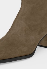 Bottine en daim marron avec un bout pointu et un talon bloc sur fond blanc.