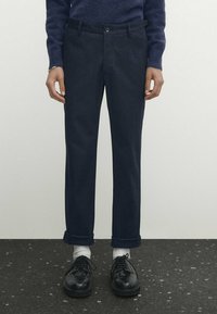 Mörkblå byxor med en slät yta, rak passform, framfickor och uppvikta ben, ihop med svarta skor och vita strumpor.