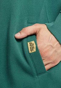 Turkusowy sweter z kieszenią z przodu, z wplecioną naszkicowaną metką, miękka faktura i akcenty w postaci ściągaczy na mankietach.