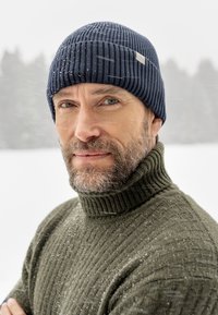 Navy ribbed knit beanie on a man wearing a dark green ribbed turtleneck sweater, with snowflakes visible against a snowy background.