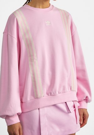 Person iført en pastelrosa Adidas sweatshirt med lodrette hvide striber og matchende rosa nederdel, stående mod en ensfarvet baggrund.