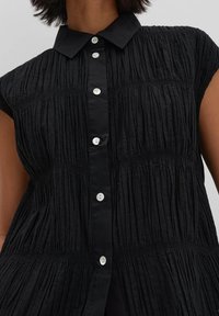 Camicetta nera a maniche corte realizzata in un tessuto texture con pieghe verticali. Presenta un colletto e una chiusura con bottoni bianchi sulla parte anteriore.