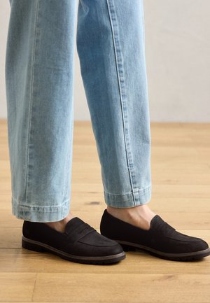 Person iført lyseblå jeans med lige ben og sorte loafers i ruskind, stående på trægulv.