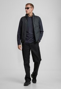 Svart quiltad väst över en marinblå långärmad tröja, kombinerat med svarta cargobyxor och svarta sneakers. Tillbehören inkluderar mörka solglasögon.