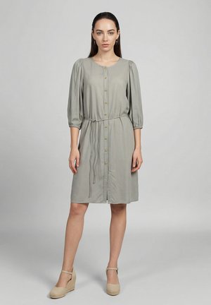 Donna che indossa un vestito midi grigio chiaro con bottoni, maniche a tre quarti gonfie e una sottile cintura in vita, abbinato a zeppe beige.