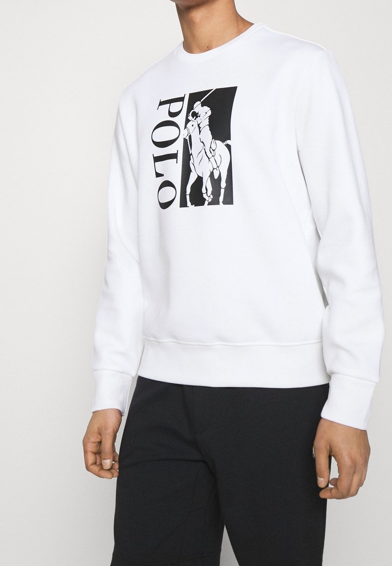 Homme portant un sweat blanc avec un logo joueur de polo noir et le mot « POLO » imprimé verticalement sur le devant, assorti à un pantalon noir.