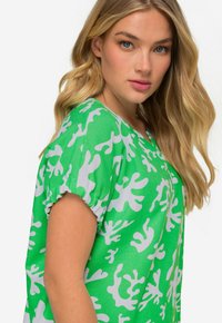 Blusa verde con motivi astratti bianchi. Maniche corte con polsini arricciati, design a V, texture di tessuto leggero.