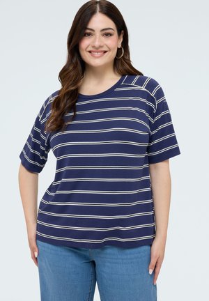 Lachende vrouw met lang bruin haar, met oorbellen in de vorm van ringen, een marineblauw-wit gestreept shirt met korte mouwen en een blauwe spijkerbroek, staand tegen een witte achtergrond.