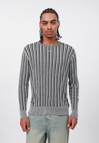 Uomo con capelli intrecciati, che indossa un maglione a righe verticali grigie e nere e jeans blu chiaro, in piedi contro uno sfondo bianco semplice.