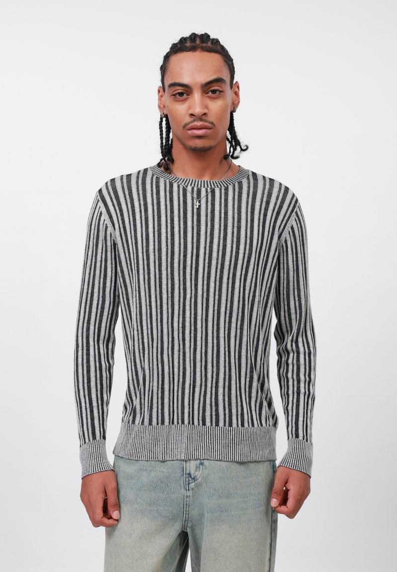 Uomo con capelli intrecciati, che indossa un maglione a righe verticali grigie e nere e jeans blu chiaro, in piedi contro uno sfondo bianco semplice.