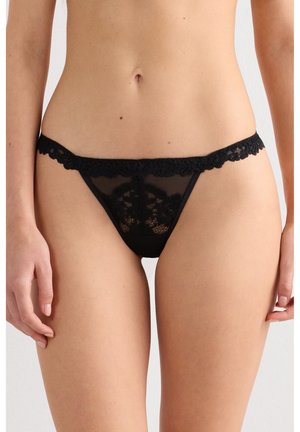 Ropa interior de bikini de encaje negro y encaje de malla, usada por una persona, mostrando la cintura frontal y los muslos superiores contra un fondo liso.