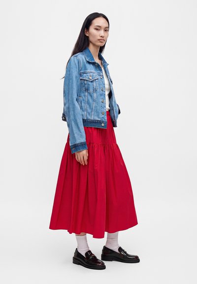 Femme aux longs cheveux foncés portant une veste en jean bleue, un top blanc, une jupe midi rouge, des chaussettes blanches et des mocassins noirs, debout sur un fond blanc.