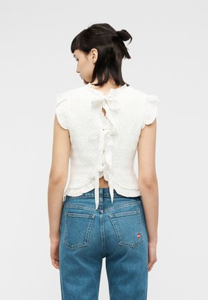 Naine, kellel on tume juuksed tagant seotud, kannab valget lameda volangidega varrukateta topi, millel on seljal lipsud, ja siniseid denimpükse.