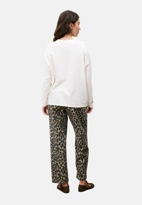 Vrouw met bruin haar, gekleed in een losse witte shirt met lange mouwen, panterprintbroek en bruine loafers, staand met de rug naar de camera op een effen achtergrond.