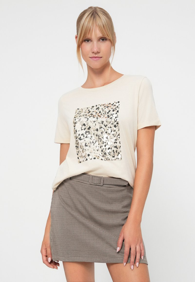 Camiseta beige con un diseño cuadrado de estampado de leopardo que presenta acentos metálicos. Combinada con una falda de cuadros marrón texturizada.