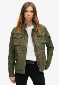 Olivenfarvet militærstil jakke med høj krave, to frontlommer og knaplukning. Har et logo på lommen.