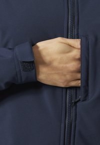 Ręka sięga do zapinanego na zamek kieszeni granatowej kurtki z rzepem przy mankiecie.