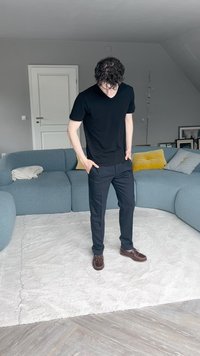 Juodas trumpas rankovės marškinėliai, tamsios kelnės ir rudos bateliai. Stovi ant šviesaus tekstilinio kilimo, matyti mėlyna audinio sofa ir pagalvėlės.