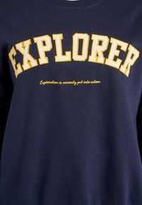 Tamsiai mėlynas megztinis su dideliu baltu ir geltonu tekstu "EXPLORER", po kuriuo yra mažesnis baltas kreivuoju šriftu parašytas šūkis. Minkšta audinio tekstūra.