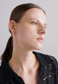 Crna čipkana košulja s cvjetnim uzorkom; glatka koža, lagani make-up, minimalni zlatni naušnice. Kosa je ravna i skupljena, neutralna pozadina.