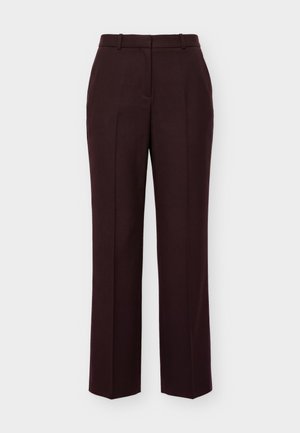Persona joven de pie, con postura recta, vistiendo una blusa negra transparente con volantes, pantalones oscuros ajustados y zapatos negros puntiagudos sobre un fondo liso.