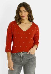 Top rojo de cuello en V con mangas tres cuartos, adornado con pequeños patrones de corazones dorados y un ribete dorado alrededor del escote. Combinado con jeans grises.