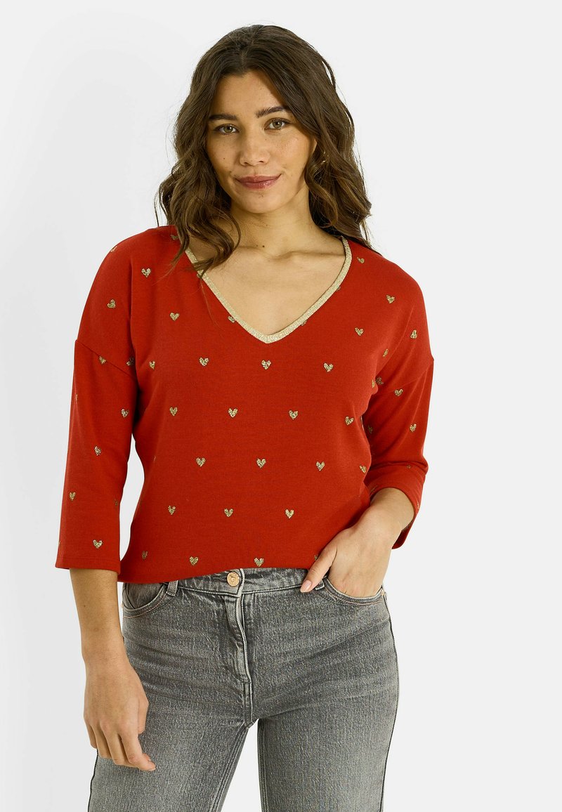 Top rojo de cuello en V con mangas tres cuartos, adornado con pequeños patrones de corazones dorados y un ribete dorado alrededor del escote. Combinado con jeans grises.