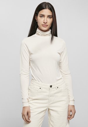 Gaiša turtlenecka bluse no gludas auduma ar garām piedurknēm, kombinēta ar gaišām korduroja biksēm ar kabatām un pogu aizdari.