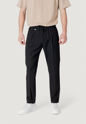 Person iført sorte slim-fit bukser med snøre i taljen og hvide sneakers, stående mod en ensfarvet lys baggrund.